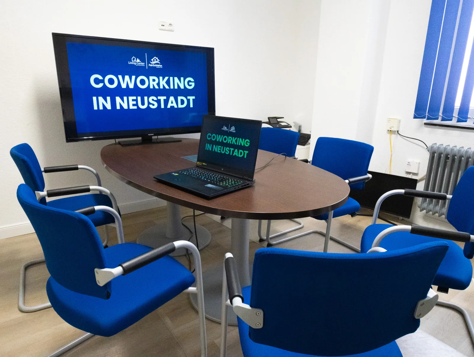 Beratungsraum mit ovalem Tisch, blauen Freischwingern und wandmontiertem Fernseher